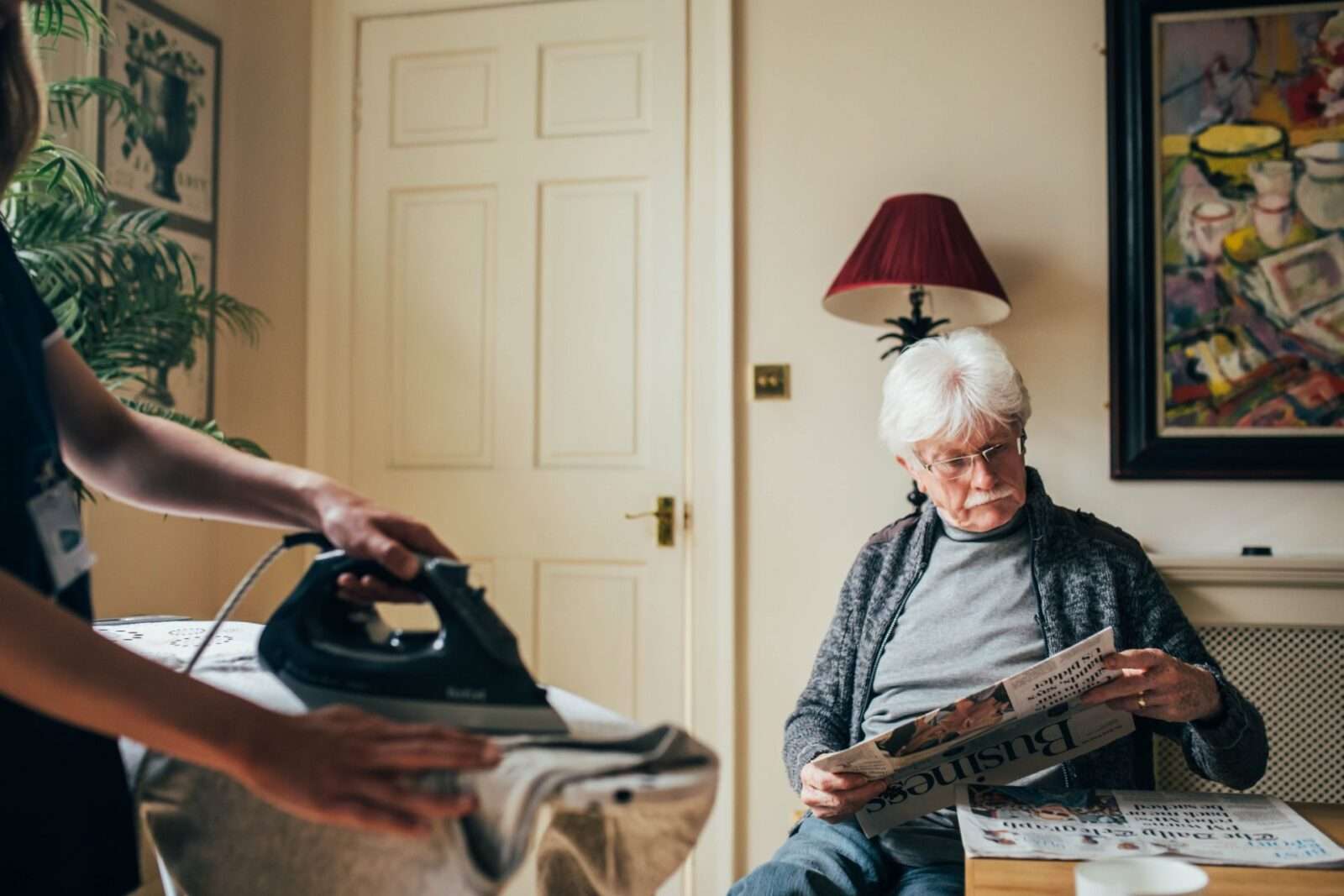 Bright Carer ironing while her client enjoys his newspaper's reading
