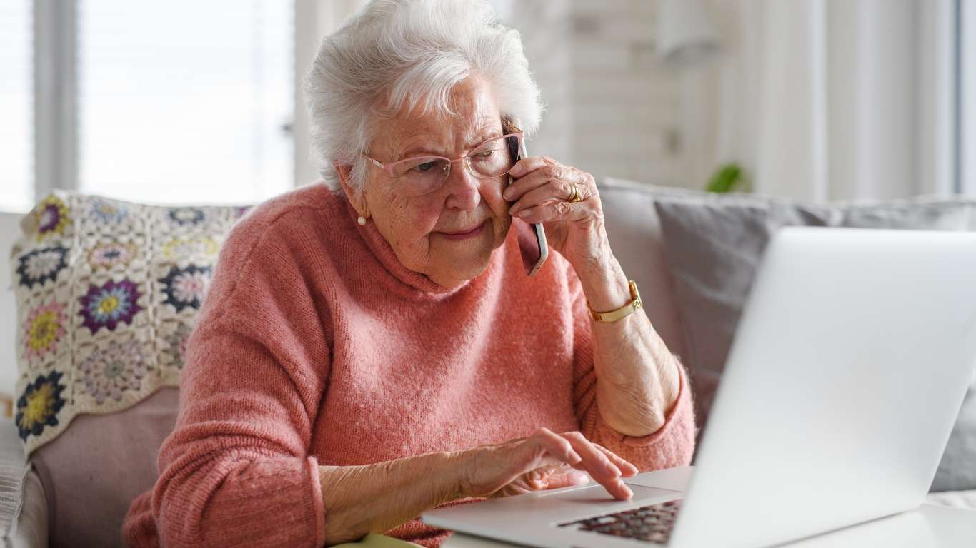 An elderly lady on the phone while using her laptop.